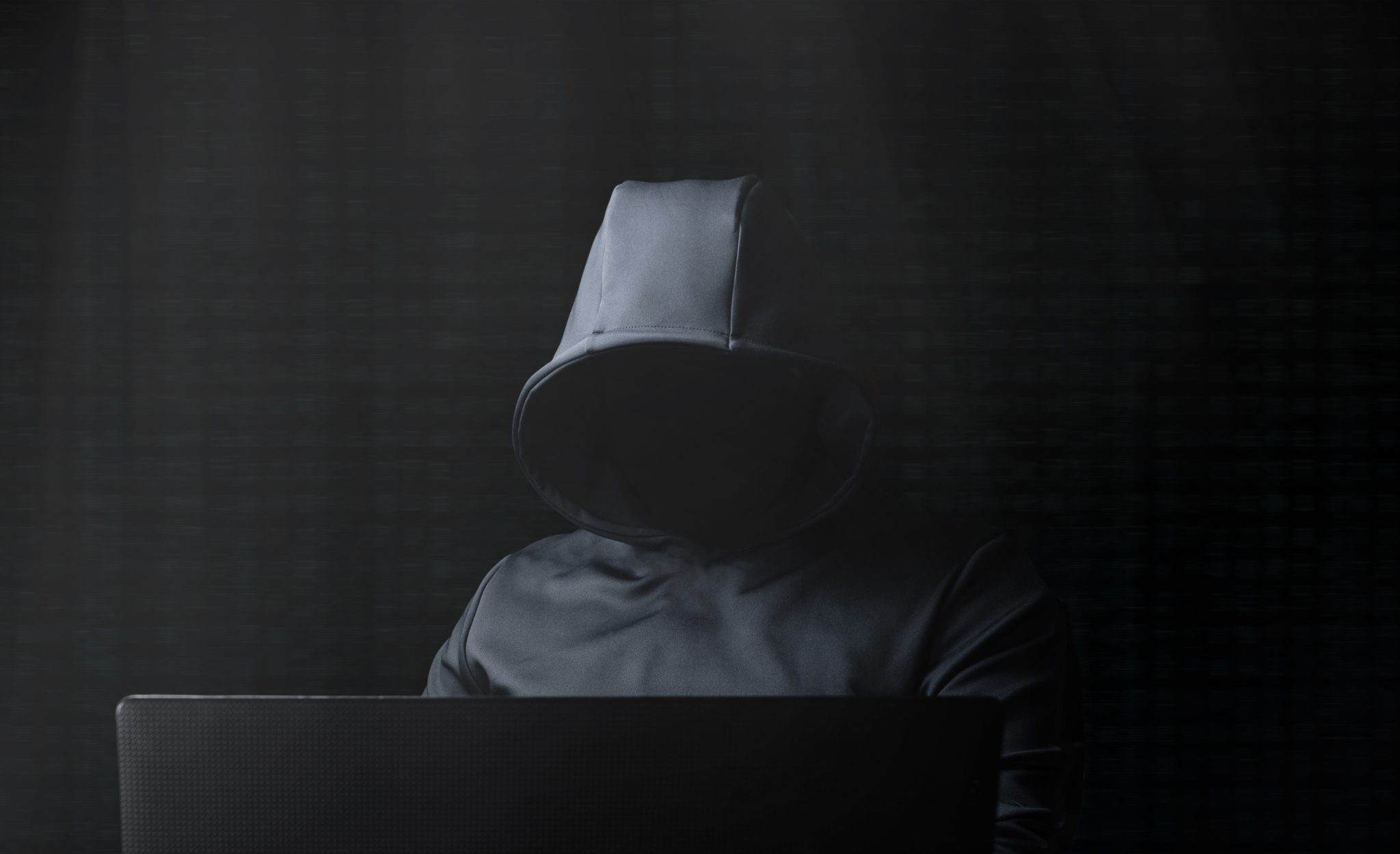 cybercriminal in the dark wearing black hoodie with face in shadows working at laptop