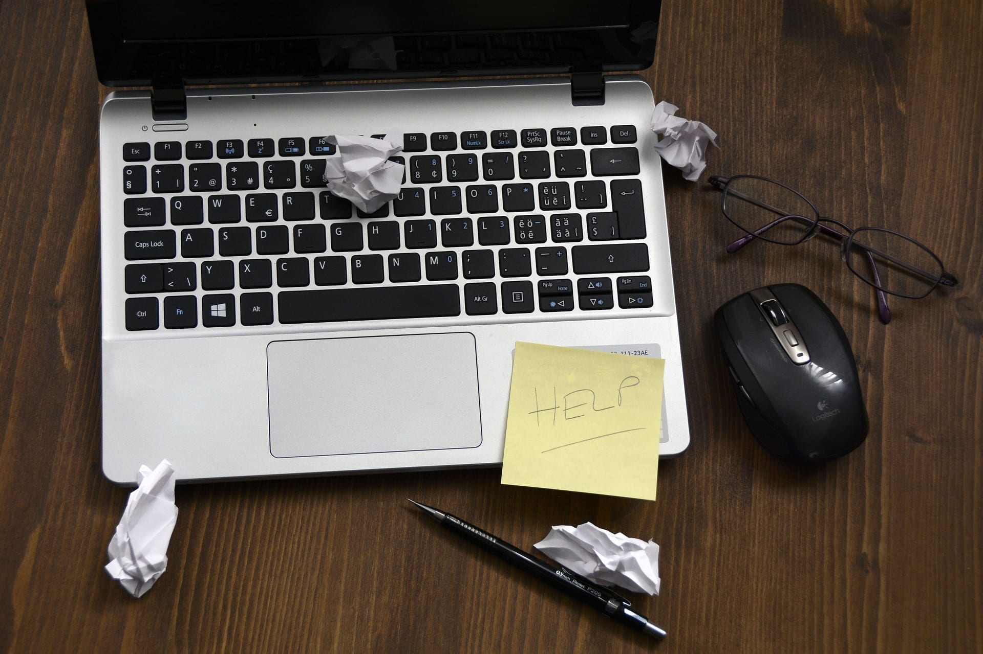 lap top open on wooden desk beside a mouse and reading glasses. Balled up papers litter the keyboard and a yellow sticky note has the word HELP written on it underlined