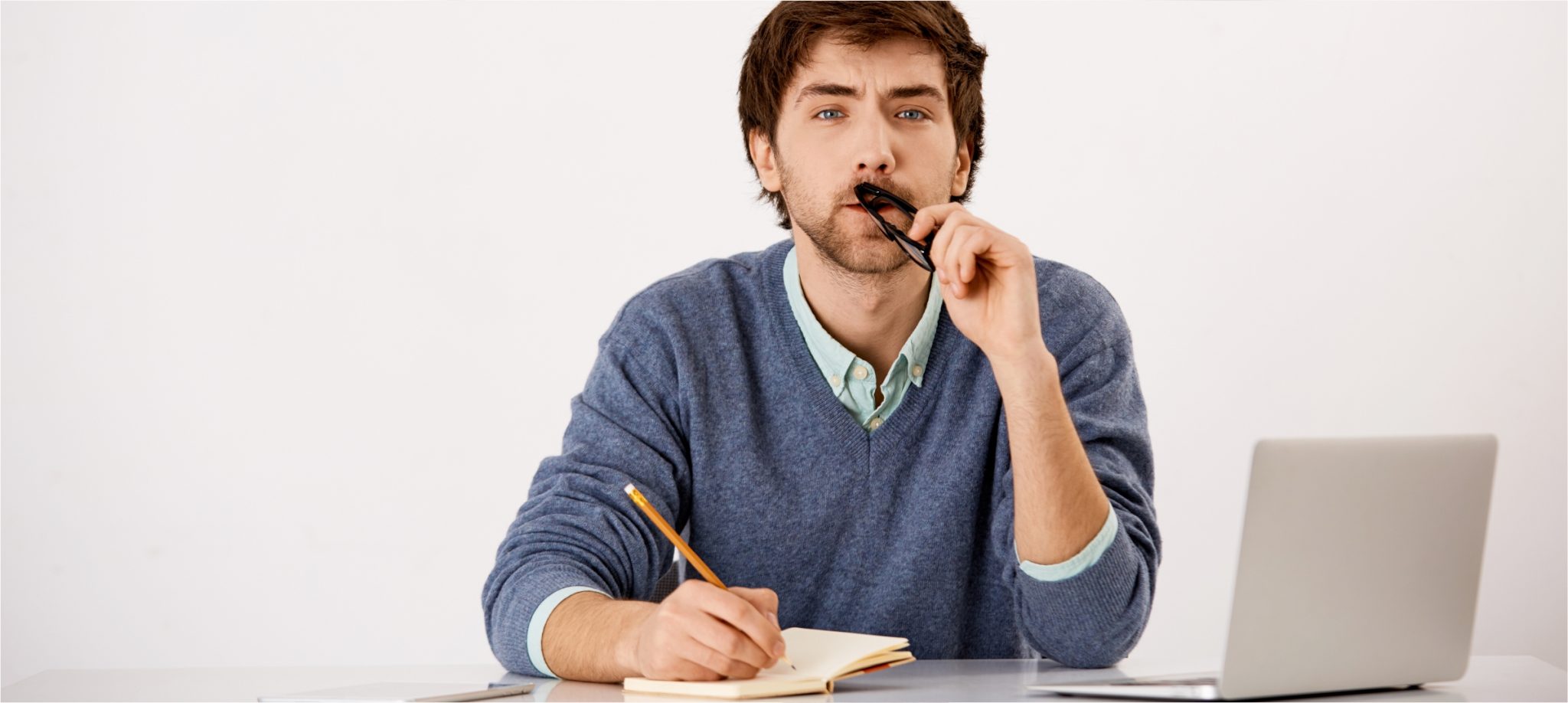 man holding pencil writing in notebook beside a laptop holding pen to mouth thinking about writing new website content