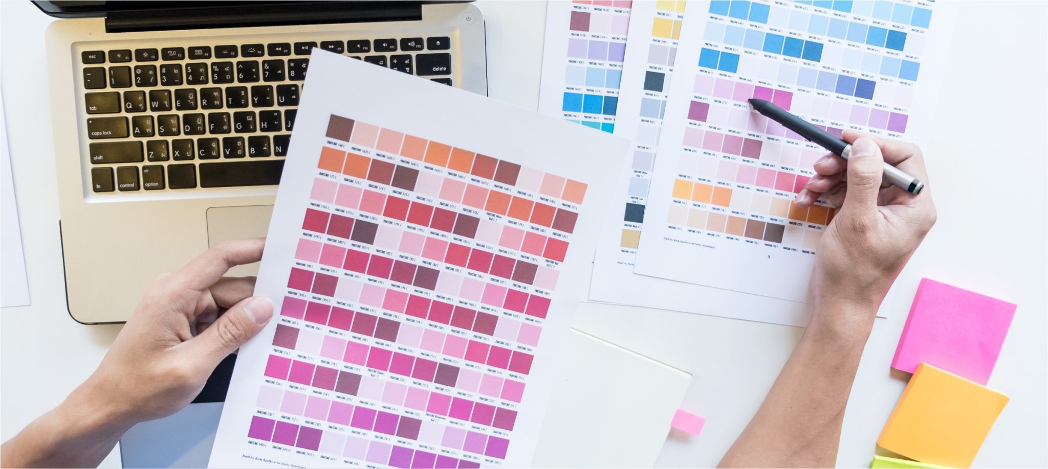 person holding page of different shades of colour over top of laptop and a pen in the other hand checking against other sheets of colours